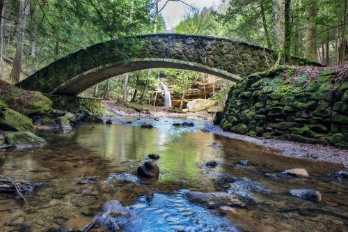 A stone arch bridge over a clear stream, surrounded by lush greenery and rocky banks in a serene forest setting.