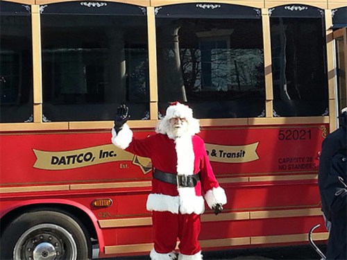 Santa Claus waves in front of a red bus with "DATCO, Inc." written on it, surrounded by trees and buildings.