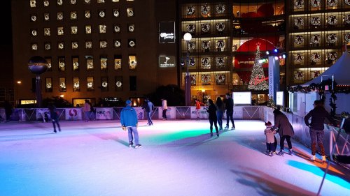 People ice skating at a festive outdoor rink, with holiday decorations and lights in the background.