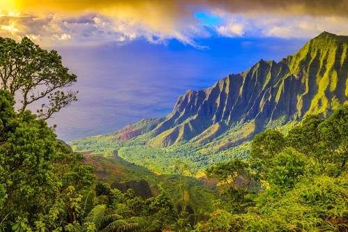 Lush green mountains rise above a serene ocean, under a dramatic sky with clouds and warm sunlight.