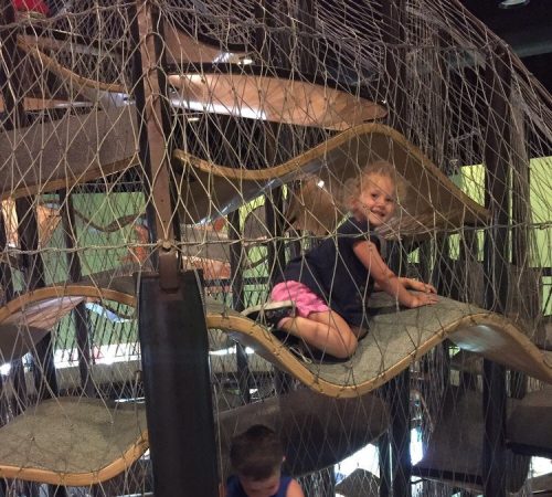 Two children play on a multi-level climbing structure with nets and wooden platforms in a play area.