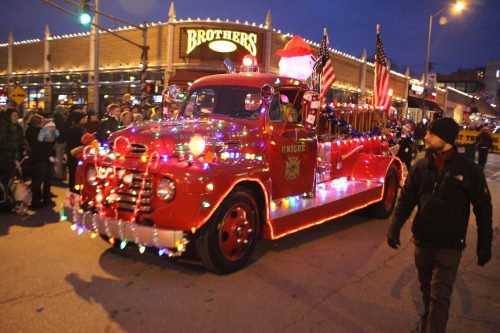 A festive red truck adorned with lights and decorations, parading through a crowd during a holiday event.