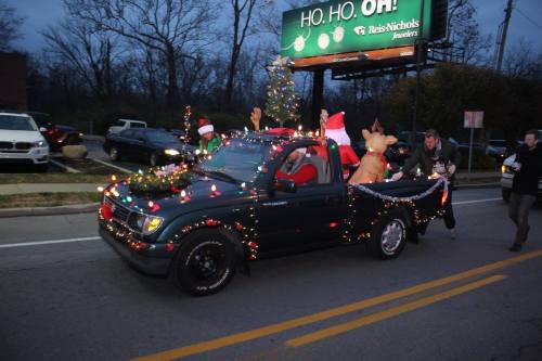 A decorated pickup truck with Christmas lights, Santa figures, and a reindeer drives down a street at dusk.