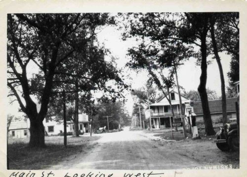 Black and white photo of a quiet street lined with trees and houses, labeled "Main St. looking west."