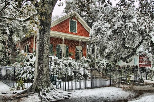 A charming red house surrounded by snow-covered trees and a white landscape, with a fence and sign in the foreground.