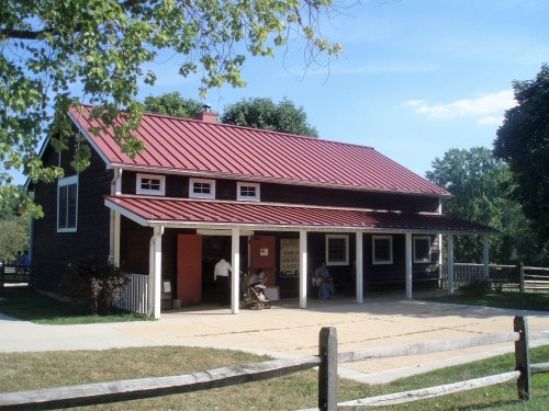 A rustic building with a red metal roof, surrounded by trees and a grassy area, featuring a porch and wooden fence.