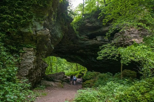 A person walks through a natural stone arch surrounded by lush green trees and rocky terrain.