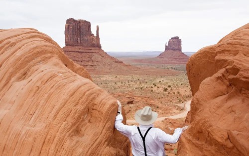 A person in a white shirt and hat stands between rock formations in a desert landscape, gazing at distant mesas.