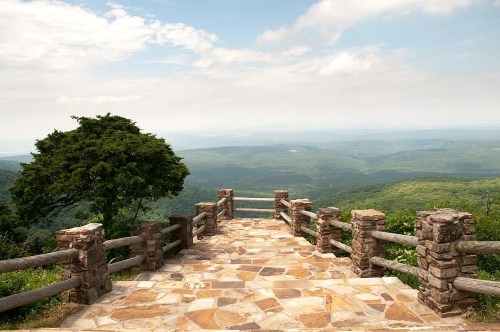 Scenic overlook with stone railing, surrounded by lush green mountains under a partly cloudy sky.