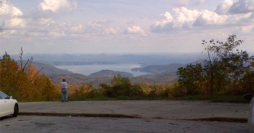 A person stands on a lookout point, gazing at a scenic view of mountains and a lake under a cloudy sky.