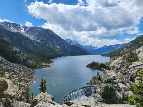 A scenic view of a lake surrounded by mountains, with a waterfall and lush greenery under a partly cloudy sky.