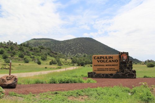 Sign for Capulin Volcano National Monument with a green landscape and mountains in the background under a cloudy sky.