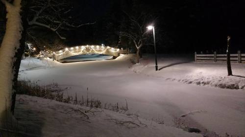 A snowy landscape at night featuring a bridge adorned with lights and a lamp post illuminating the scene.
