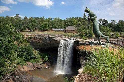 A statue of a Native American figure overlooks a waterfall surrounded by lush greenery and a clear blue sky.