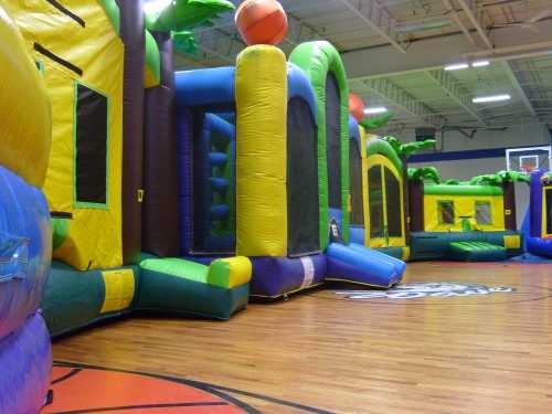 Colorful inflatable bounce houses and slides in a gymnasium with a wooden floor.