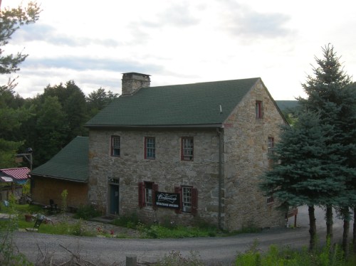 A rustic stone house with a green roof, surrounded by trees and a gravel path, under a cloudy sky.