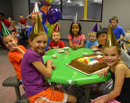 A group of children wearing party hats smiles around a table with a colorful cake and decorations.