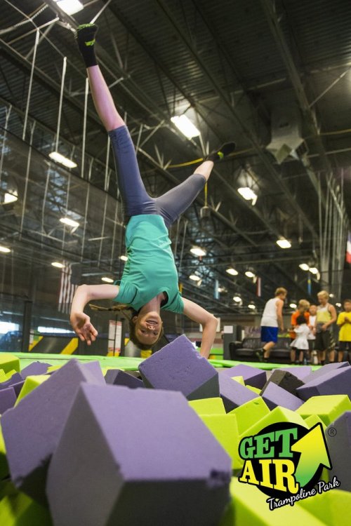 A girl performs a handstand above colorful foam blocks at a trampoline park, surrounded by other jumpers.