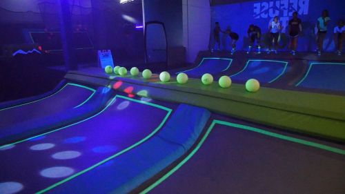 A vibrant indoor trampoline park with neon lights and green outlines, featuring a row of glowing balls on a ramp.