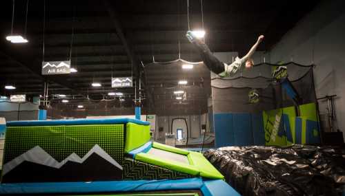 A person jumps off a trampoline, soaring through the air above a colorful landing area in a trampoline park.