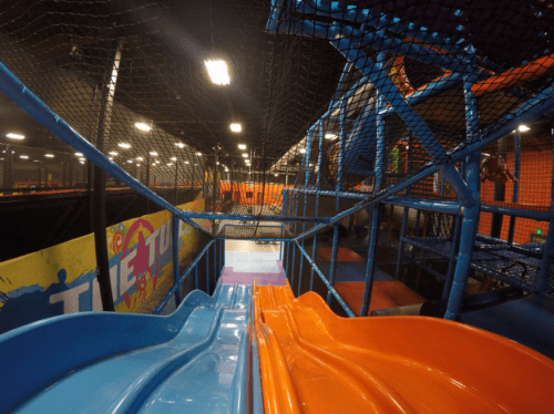 View of colorful slides in a trampoline park, surrounded by safety nets and vibrant play areas.