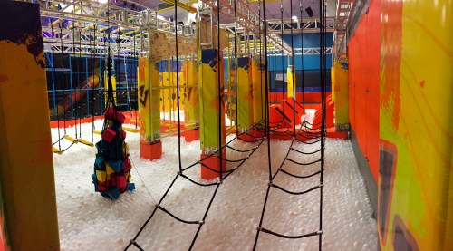 A colorful indoor play area with climbing structures and a foam pit, featuring ropes and vibrant walls.