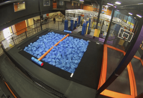 A trampoline park featuring a large foam pit filled with blue foam blocks and an orange balance beam on top.