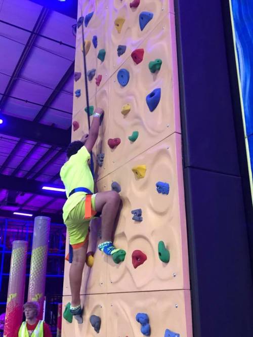 A child climbs a colorful indoor rock wall, wearing bright shorts and shoes, with a safety harness attached.