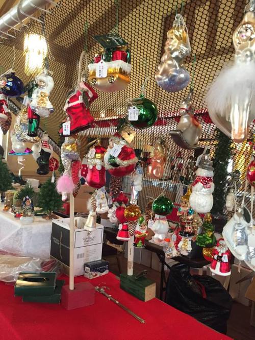 A festive display of colorful Christmas ornaments hanging on a net, with small trees and a red tablecloth in the foreground.