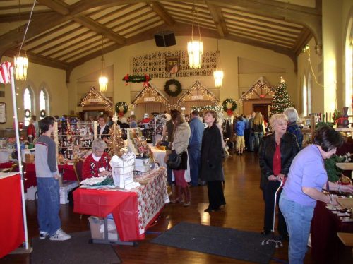 A bustling holiday market with vendors, shoppers, and festive decorations in a large, warmly lit hall.