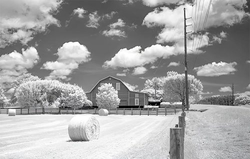 A serene rural landscape featuring a barn, hay bales, trees, and a cloudy sky, captured in infrared.