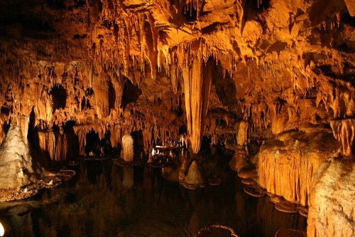 A stunning underground cave with stalactites and stalagmites reflected in a calm water pool, illuminated by warm light.