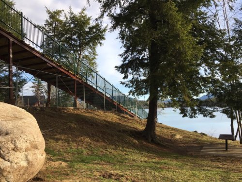 A sloped pathway leads to a lake, surrounded by trees and a rocky area, with a fence along the path.