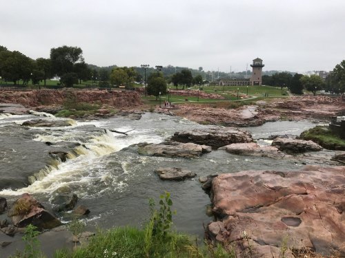A scenic view of a river with rocky banks, surrounded by greenery and a distant tower on a cloudy day.