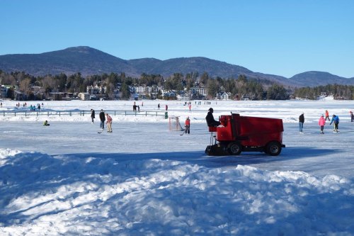 A red ice resurfacer on a snowy rink with people skating and playing hockey in the background and mountains in the distance.