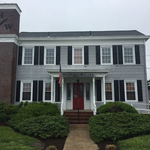 A gray building with black shutters and a red door, featuring the sign "Harpoon Willy's" and an American flag.