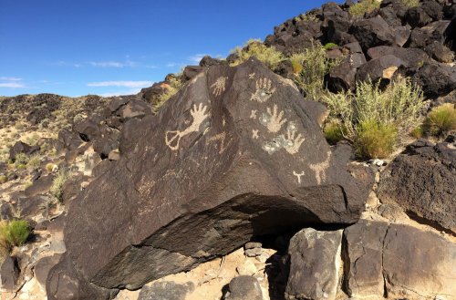 Petroglyphs featuring handprints and symbols on a large rock in a desert landscape.