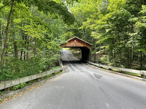 A wooden covered bridge spans a road, surrounded by lush green trees and dappled sunlight.