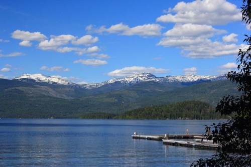 A serene lake surrounded by mountains, with snow-capped peaks and fluffy clouds in a clear blue sky.