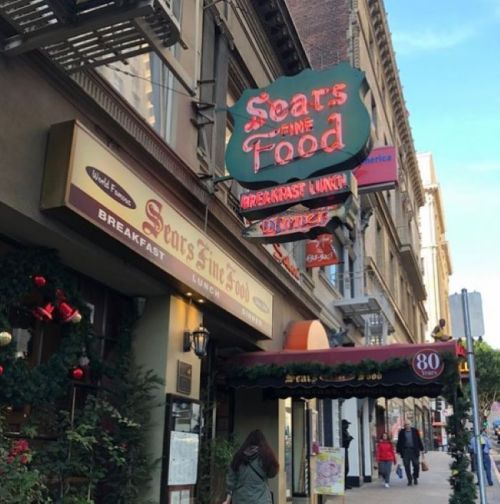 Sign for "Sears Fine Food" with a vintage design, featuring "Breakfast" and "Lunch" in a bustling city setting.