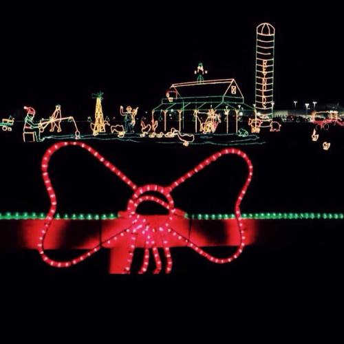 A festive display of holiday lights featuring a large red bow in the foreground and a decorated barn in the background.