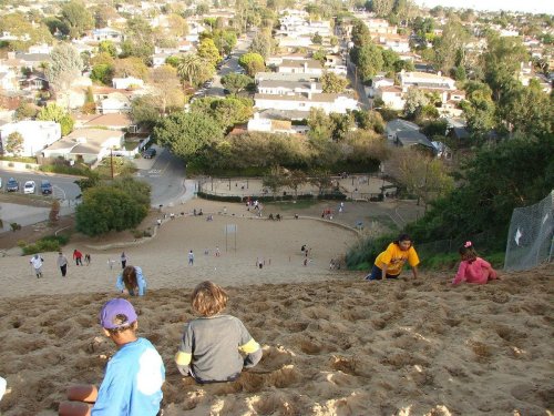 A sandy hill with children climbing and playing, overlooking a neighborhood with houses and a park below.