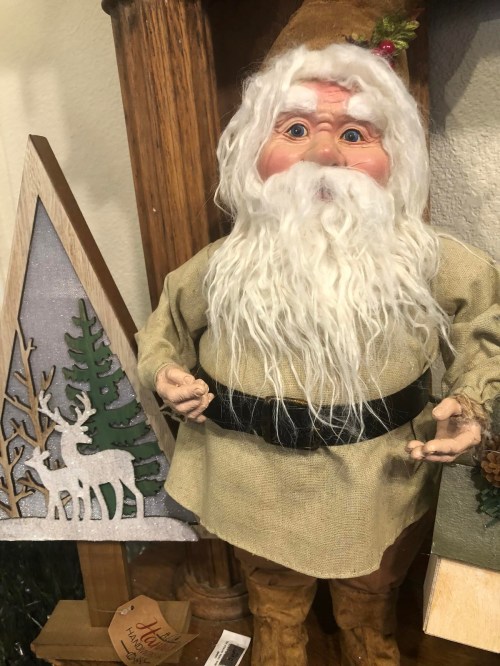 A decorative figure with a long white beard, wearing a beige outfit and black belt, stands beside a wooden Christmas tree.