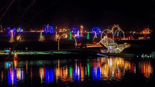 Colorful holiday lights illuminate a park at night, reflecting on a calm water surface.