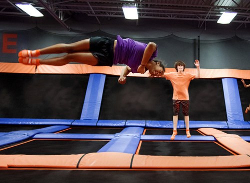 A child jumps high on a trampoline, while another child watches nearby in a trampoline park.
