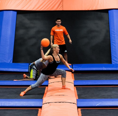 A woman jumps on a trampoline, throwing a ball, while a man watches in the background. Bright orange and blue setting.