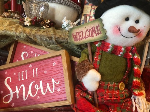 A cheerful snowman in a plaid outfit holds a "Welcome" sign, surrounded by festive decorations and signs saying "Let It Snow."