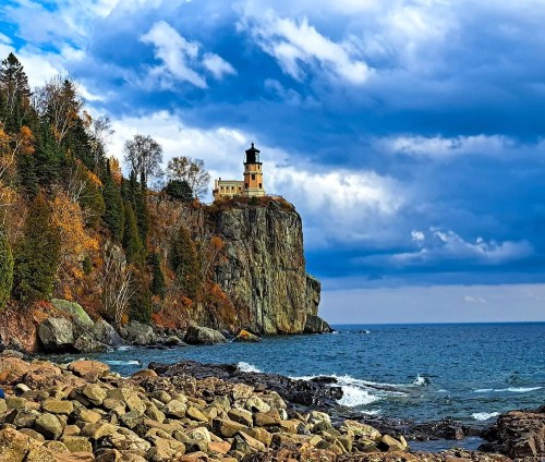 A lighthouse perched on a rocky cliff, surrounded by autumn trees and a cloudy sky over the ocean.