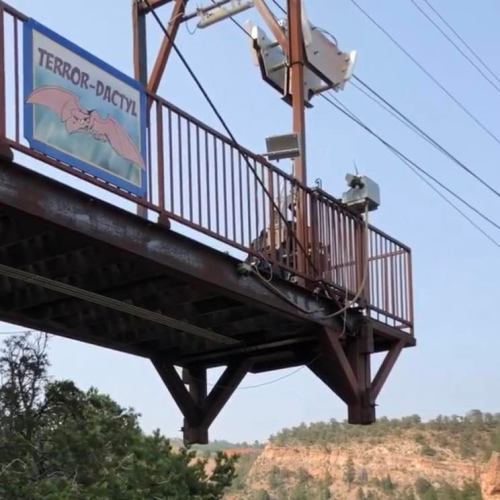 A metal bridge with a sign reading "TERROR-DACTYL" and surveillance cameras, overlooking a scenic landscape.