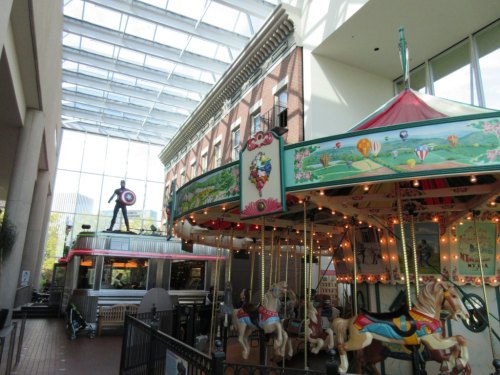 A colorful carousel inside a mall, with a glass ceiling and a statue of a superhero in the background.